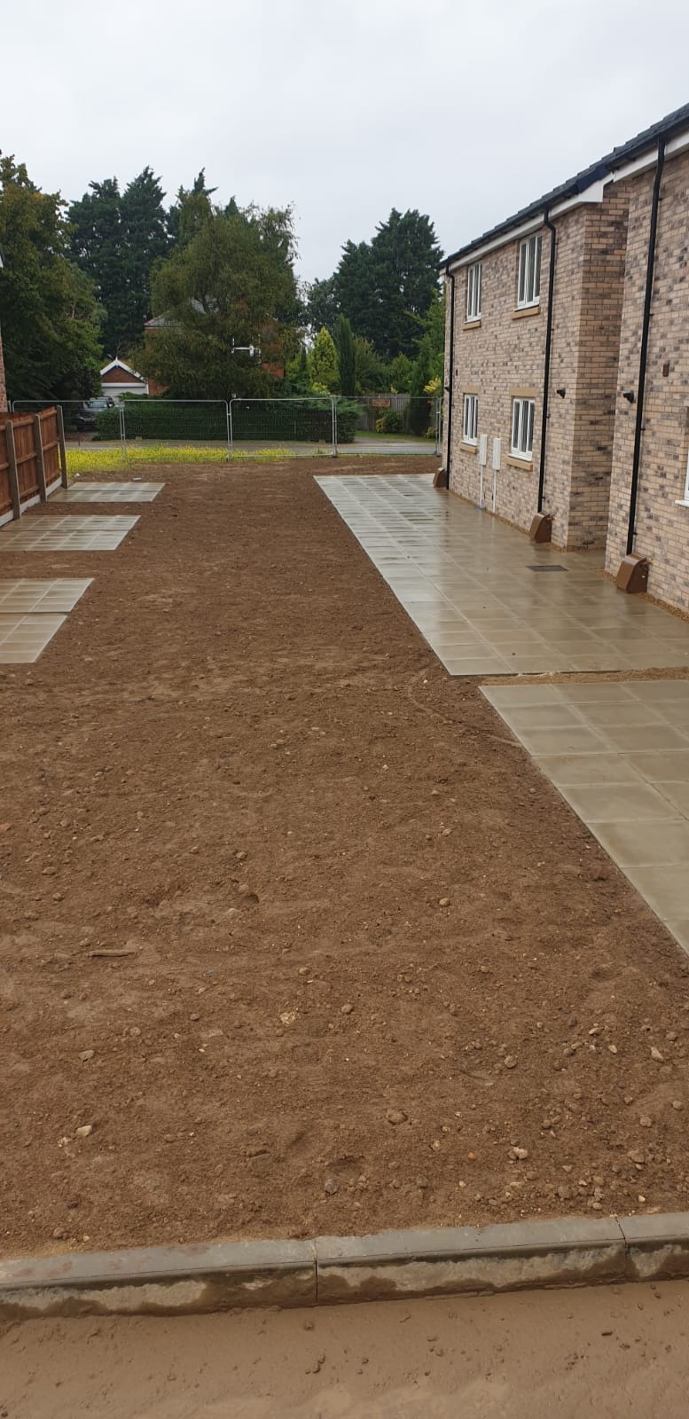 Przygotowanie terenu pod przyszły chodnik z płyt betonowych, widoczne fragmenty ułożonych płyt oraz wyrównana ziemia, w tle budynek mieszkalny i ogrodzenie.