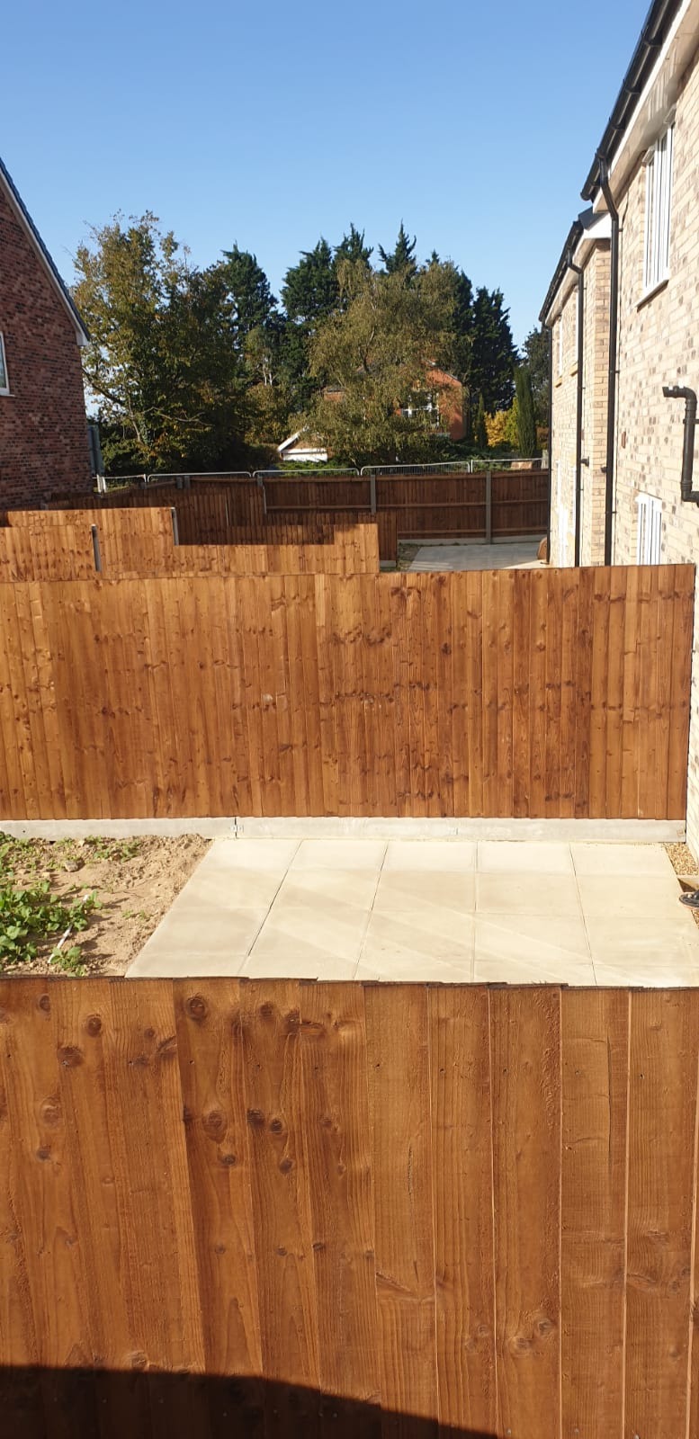 Nowo postawione, drewniane ogrodzenie panelowe w kolorze brązowym, oddzielające posesje, z widocznymi płytami chodnikowymi ułożonymi wzdłuż ogrodzenia i fragmentami zieleni w tle.