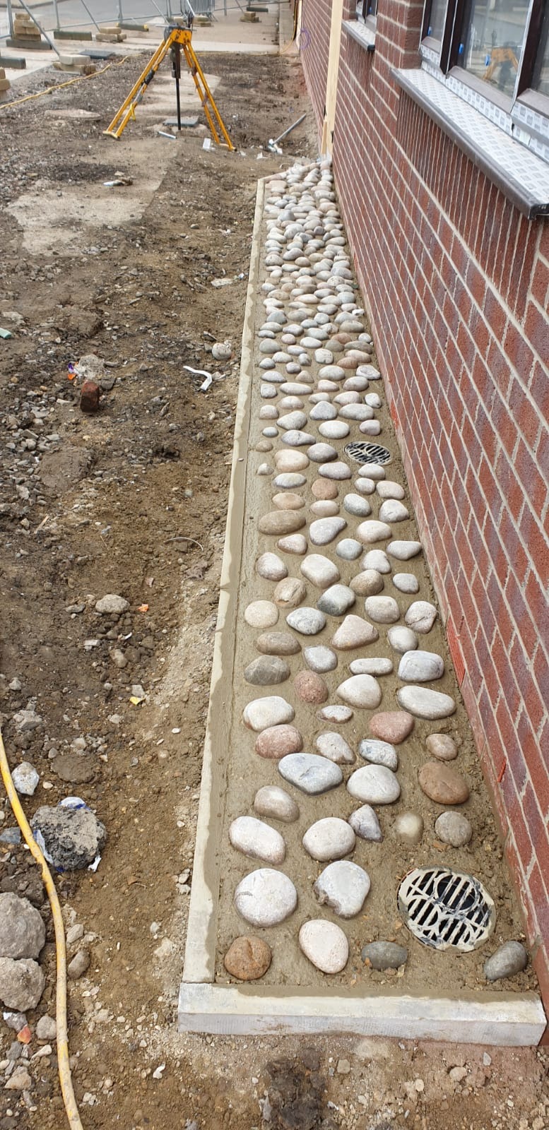 Wykończenie obrzeża budynku w Peterborough za pomocą betonowej ramy i dekoracyjnych, otoczaków rzecznych w jasnych odcieniach szarości i beżu, z kratką odpływową w narożniku.