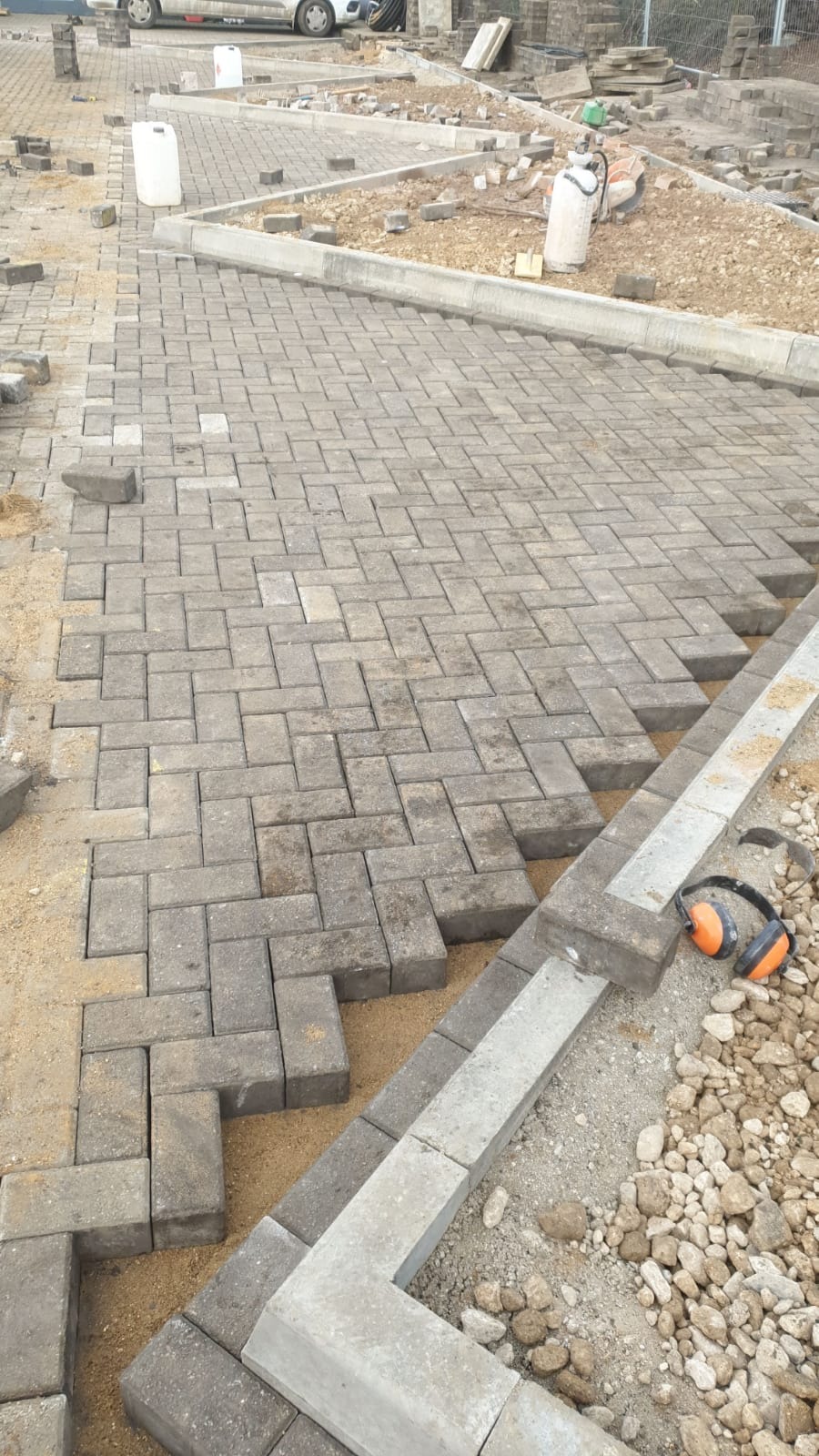 Fragment układanej nawierzchni z szarej kostki brukowej, częściowo zasypanej piaskiem, z widocznymi obrzeżami i roboczymi słuchawkami ochronnymi w prawym dolnym rogu.