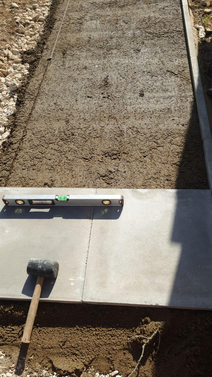 Wyrównywanie podłoża pod kostkę brukową z użyciem poziomicy i gumowego młotka, widoczne obrzeża i podsypka z kruszywa.