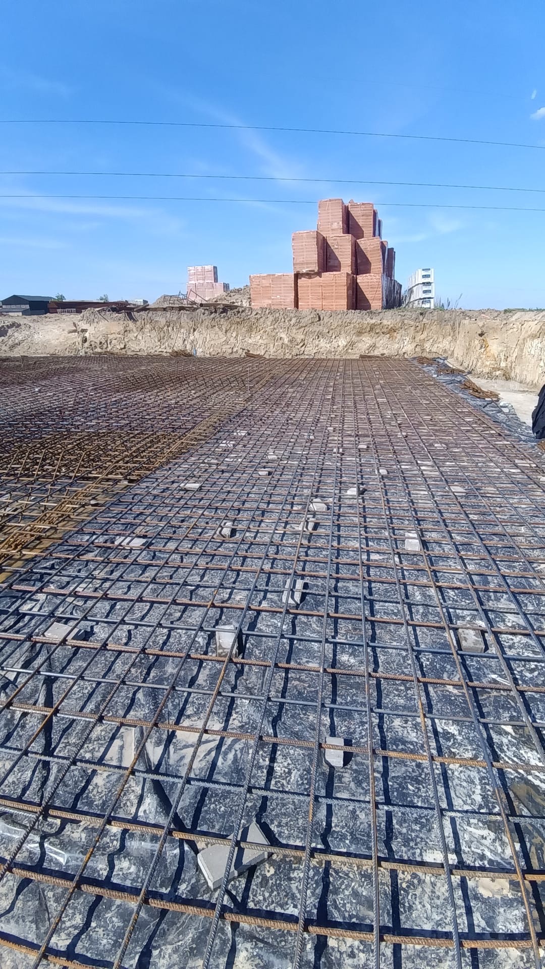 Siatka zbrojeniowa na folii, przygotowana do zalania betonem. W tle cegły i wykop pod fundamenty, błękitne niebo.
