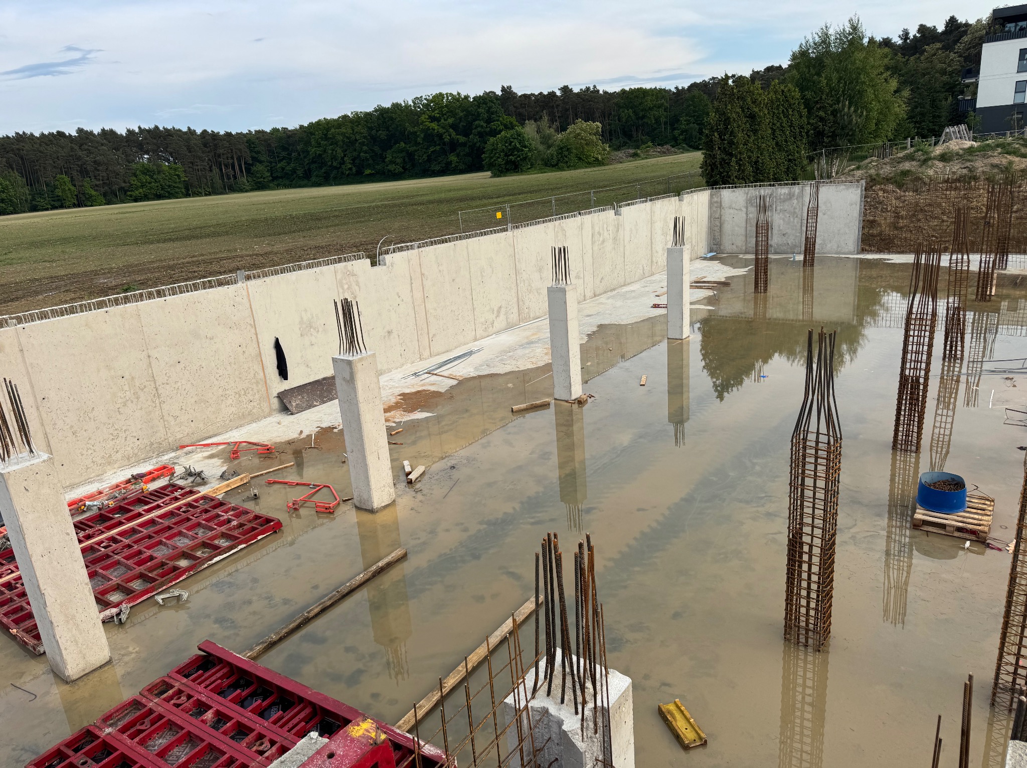 Zalane fundamenty z wystającymi zbrojeniami pionowymi i betonowymi słupami. W tle betonowa ściana oporowa i zielone drzewa. Widoczne czerwone szalunki.