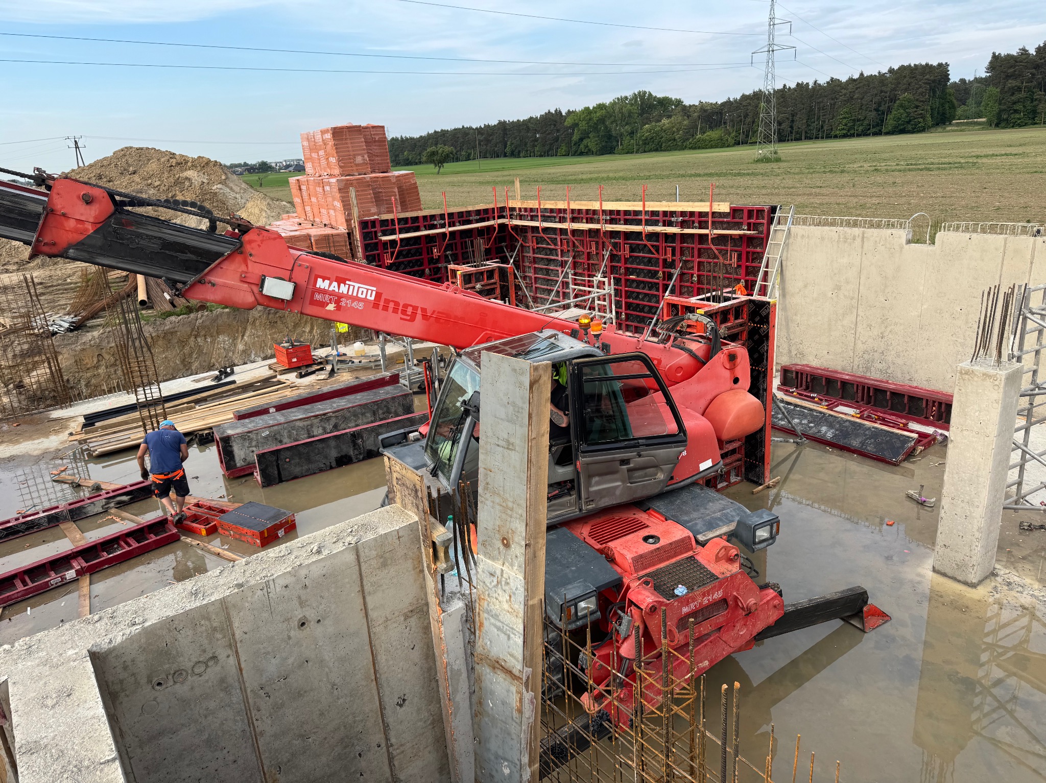Budowa żelbetowej konstrukcji z użyciem czerwonego dźwigu Manitou. Widoczne szalunki, zbrojenie i częściowo zalane fundamenty. W tle drzewa i słupy wysokiego napięcia.