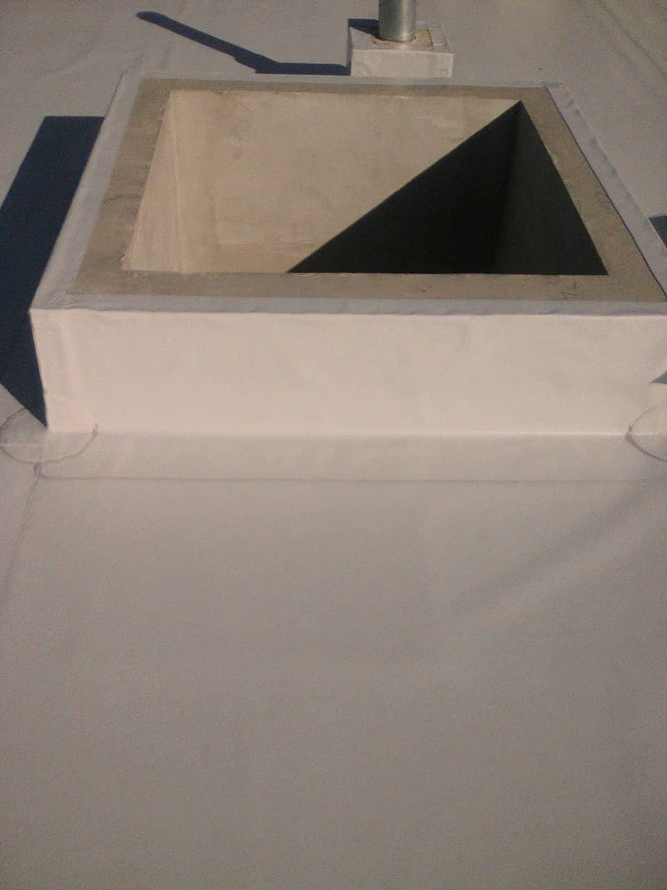 Szczegółowe ujęcie płaskiego dachu z wykończonym kominem wentylacyjnym w kształcie kwadratu, z widocznym cieniem wewnątrz komina i na powierzchni dachu.