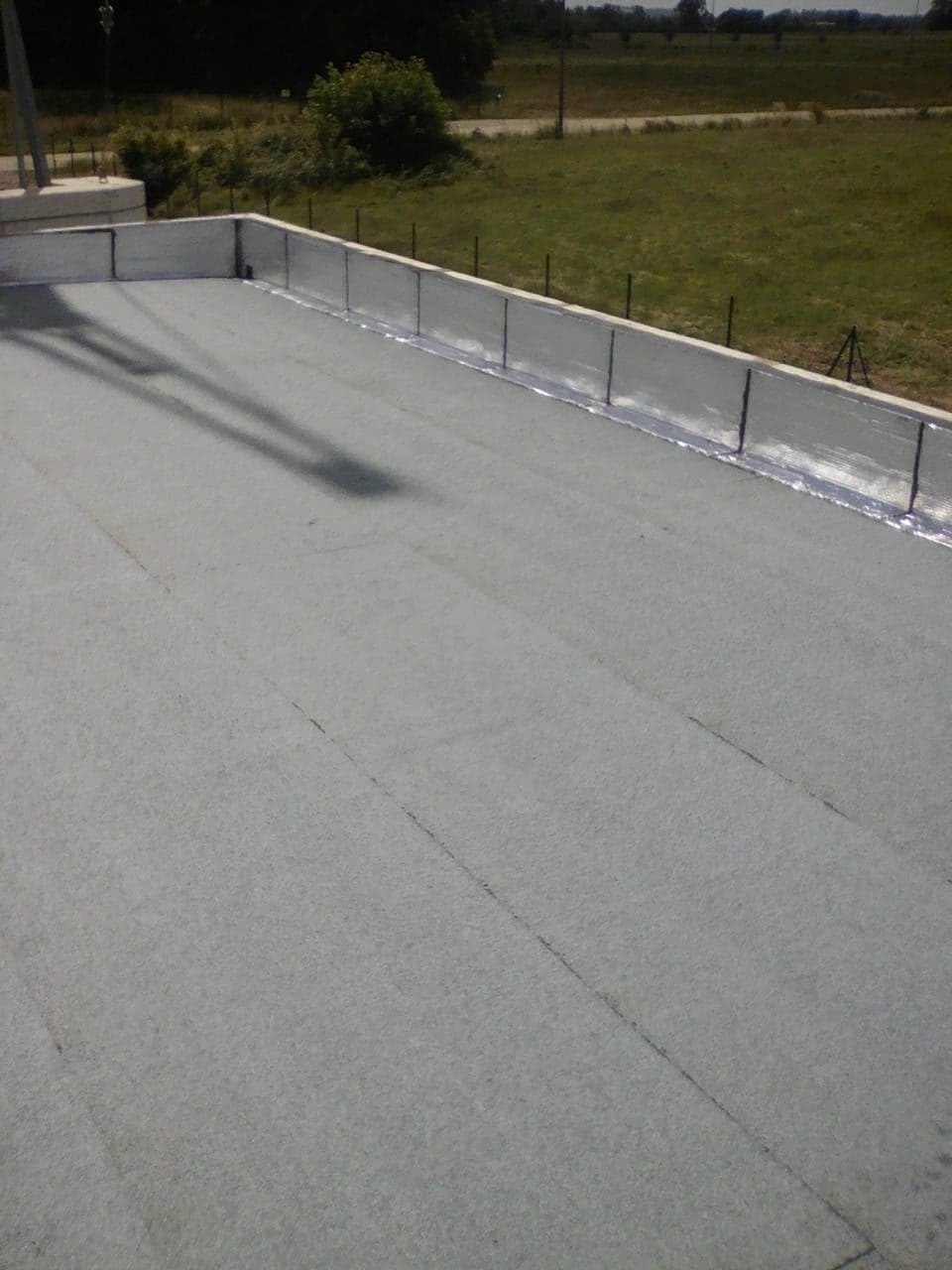 Płaski dach pokryty szarą papą termozgrzewalną, z widocznymi łączeniami brytów i obróbkami blacharskimi przy attyce, na tle zielonego krajobrazu.