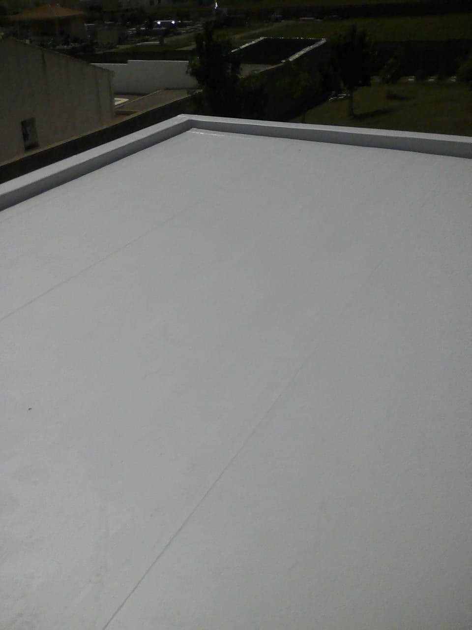 Szary, płaski dach z widocznymi liniami łączeń, otoczony niskim obramowaniem, z budynkami i zielenią w tle.