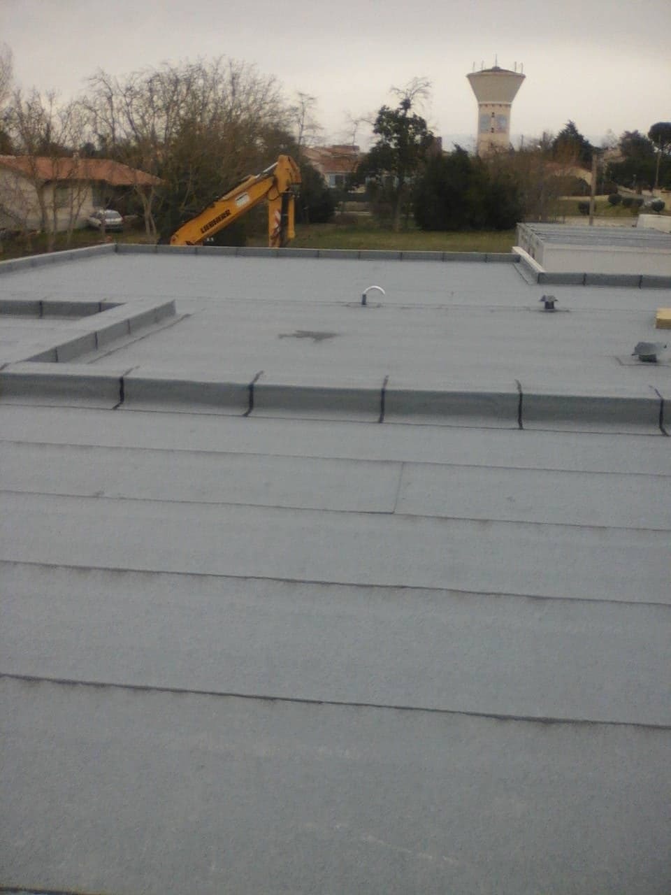 Płaski dach pokryty szarą papą, widoczny fragment z kominkiem wentylacyjnym, w tle żółta koparka i wieża ciśnień.