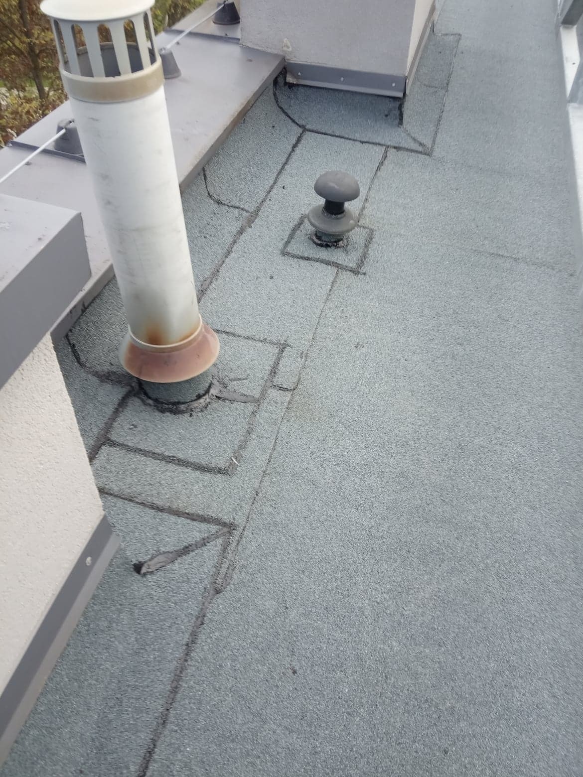 Płaski dach pokryty papą z widocznymi kominkami wentylacyjnymi i obróbkami blacharskimi, uszczelniony czarnym kitem dekarskim.