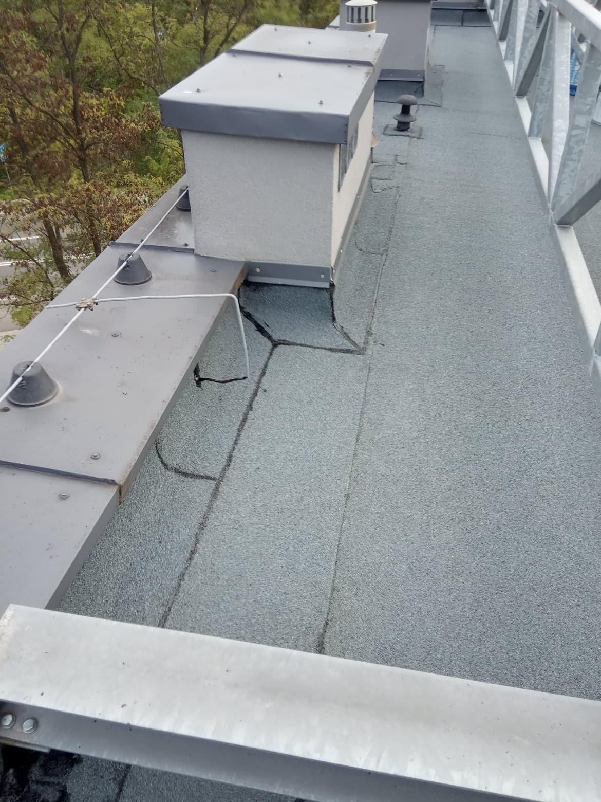 Szary, płaski dach pokryty papą termozgrzewalną z widocznymi kominami wentylacyjnymi i elementami zabezpieczającymi przed upadkiem z wysokości.