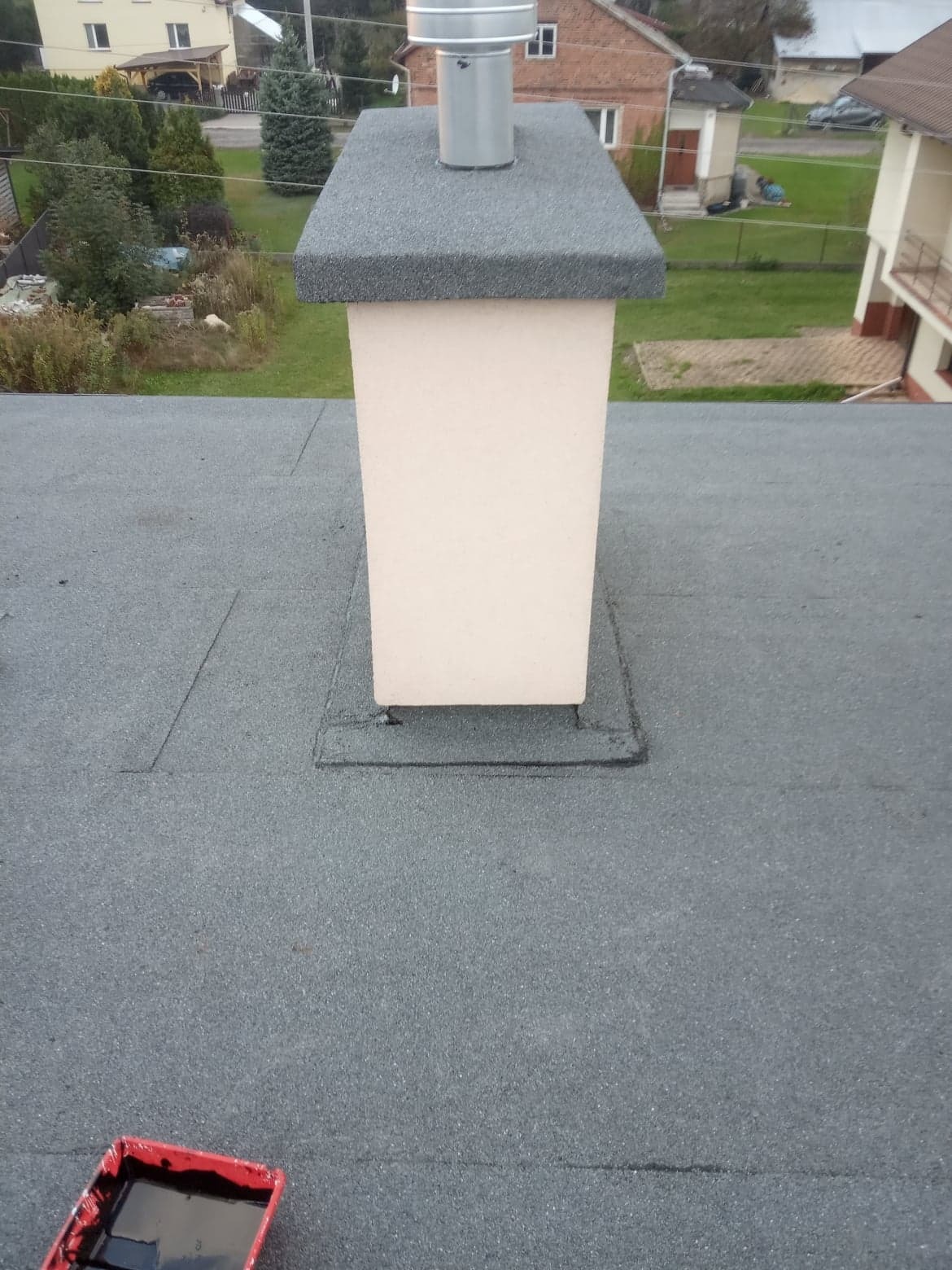 Widok z góry na dach pokryty papą bitumiczną z kominem i pojemnikiem z czarną substancją uszczelniającą