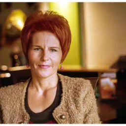 Portret kobiety z krótkimi rudymi włosami, ubranej w beżową kurtkę, z kolczykami w kształcie kulek, siedzącej w pomieszczeniu o ciepłym oświetleniu.