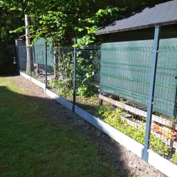 Art-Clean - Nowo zamontowane, panelowe ogrodzenie z podmurówką betonową, siatka w kolorze grafitowym, widok z perspektywy trawnika, w tle zieleń drzew i budynek gospodarczy z siatką maskującą.