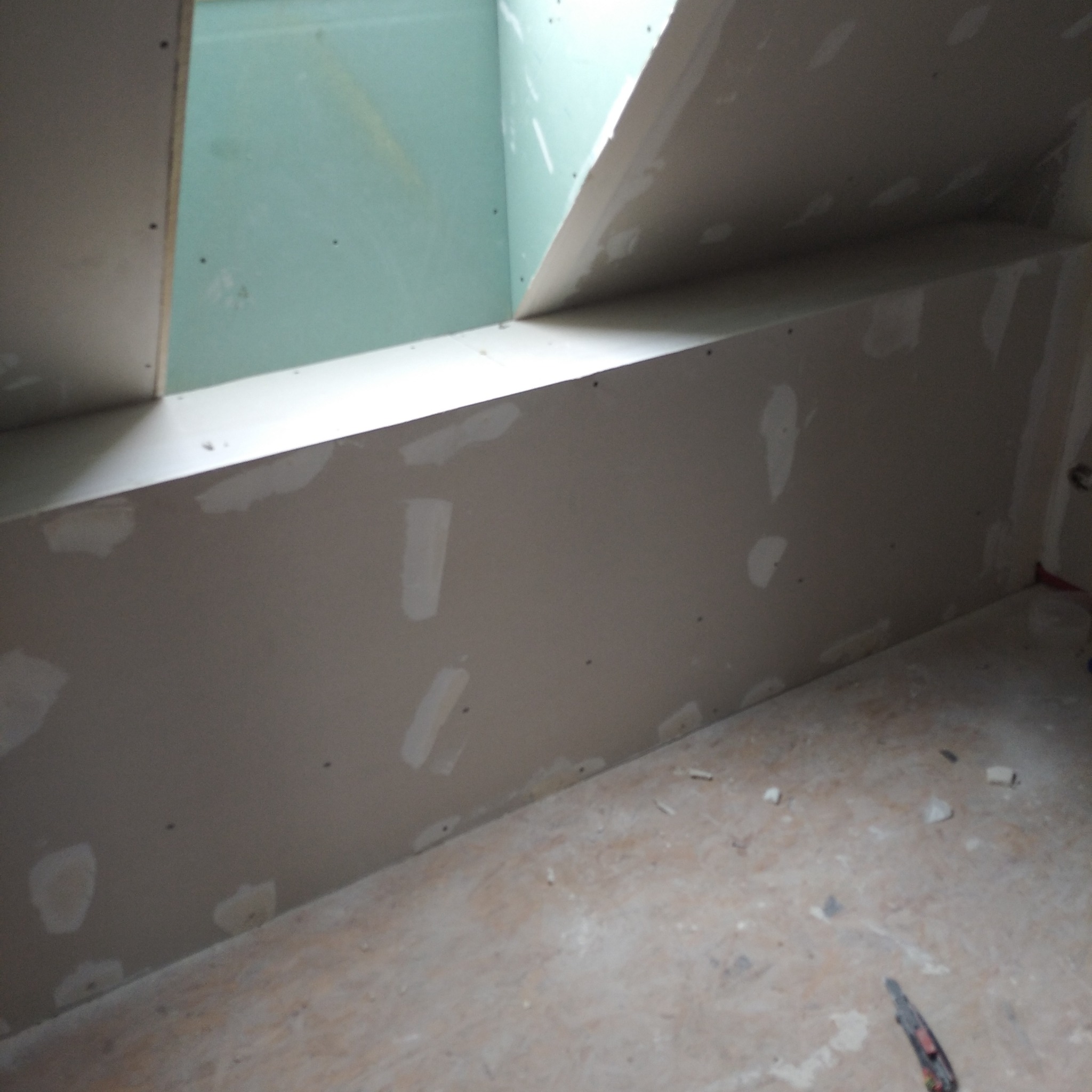 Wnętrze w trakcie wykańczania: ściany z zamontowaną płytą gipsowo-kartonową, wstępnie wyszpachlowane, widoczne łączenia i śruby, podłoga z resztkami materiałów budowlanych, częściowo widoczny nóż...