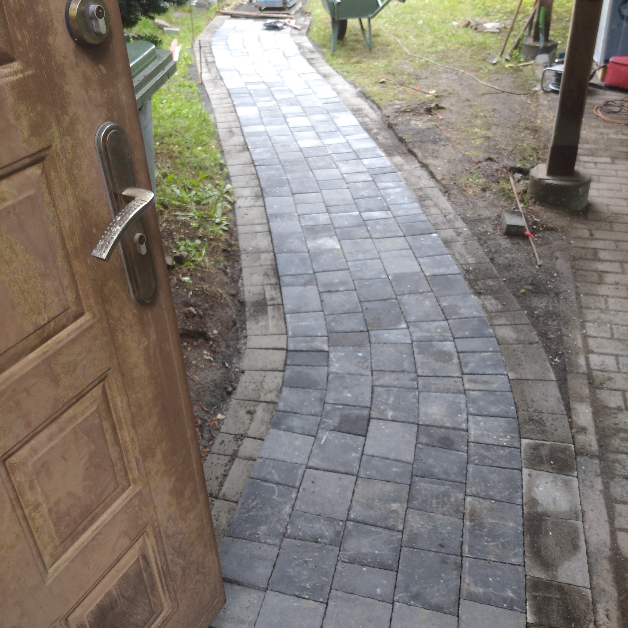 Świeżo ułożony, wijący się chodnik z szarej kostki brukowej w Legnicy, widziany zza uchylonych, drewnianych drzwi wejściowych.