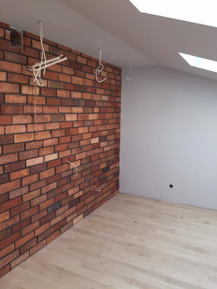 Wnętrze poddasza z fragmentem ściany wyłożonej cegłą klinkierową, jasną drewnianą podłogą i pomalowaną na szaro ścianą, widoczne kable wystające ze ściany, okno dachowe.