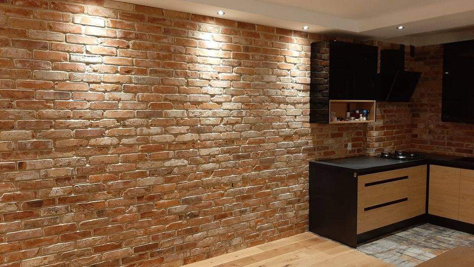 Ściana z cegły dekoracyjnej w kuchni z nowoczesnymi czarnymi szafkami i drewnianymi frontami, oświetlona punktowo, z widocznymi fugami i nieregularną fakturą cegieł.