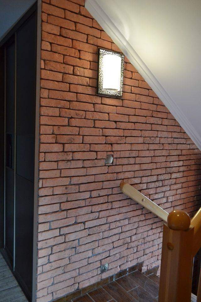 Ściana z cegły klinkierowej z oświetleniem punktowym w dekoracyjnej oprawie, widoczny fragment schodów z drewnianą balustradą i ciemna szafa wnękowa.