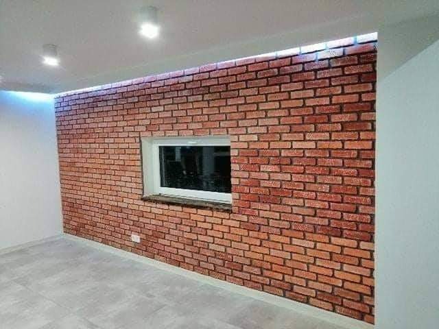 Ściana wewnętrzna wyłożona czerwoną cegłą z oknem, widoczne szare panele podłogowe i biały sufit z oświetleniem punktowym.