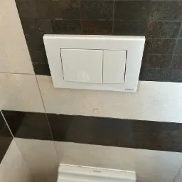 Biały panel spłuczki toalety firmy Valsir zamontowany na ścianie z ciemnobrązowych i beżowych płytek.