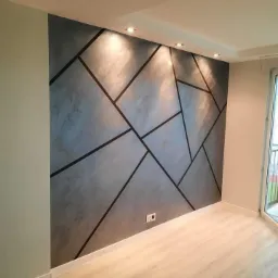 Ściana z dekoracyjną szarą strukturą i geometrycznym wzorem czarnych linii, oświetlona punktowymi lampami sufitowymi, widoczny fragment parkietu i okna balkonowego.