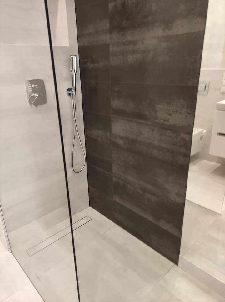 Nowoczesna kabina prysznicowa z szarymi płytkami imitującymi rdzę na ścianie, szklana ścianka z czarnym wykończeniem i odpływ liniowy w podłodze.