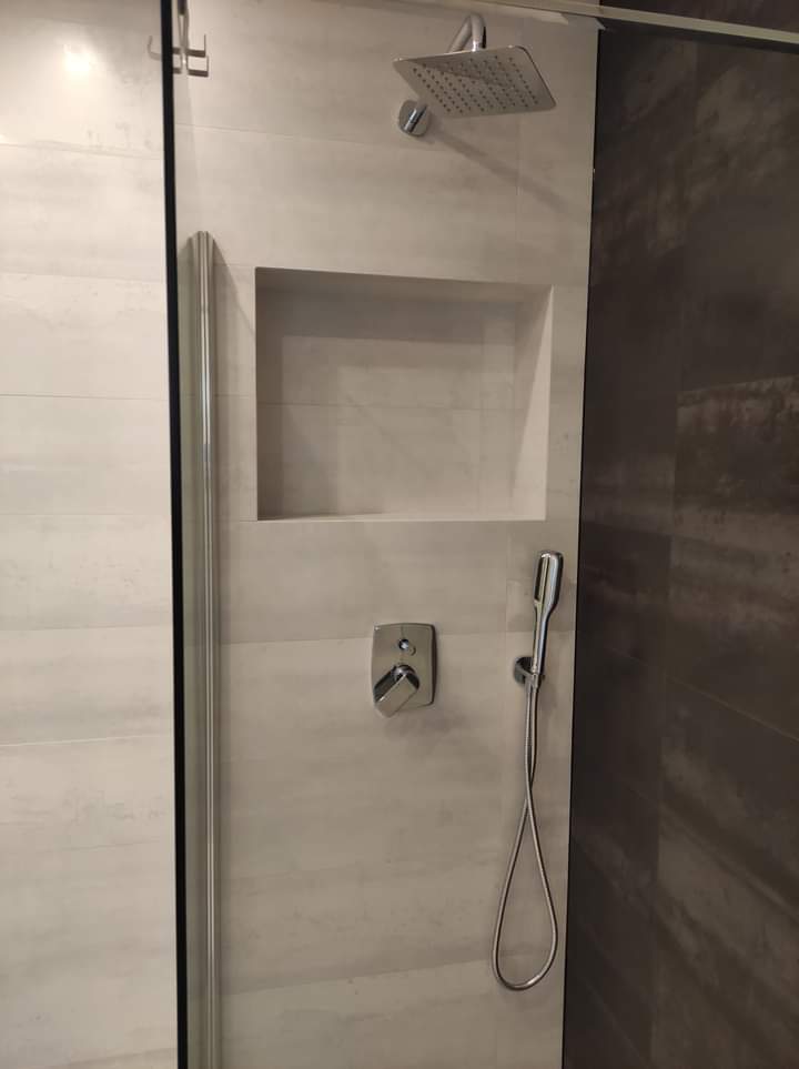 Nowoczesna kabina prysznicowa z szarymi płytkami, wnęką na kosmetyki, deszczownicą i słuchawką prysznicową.