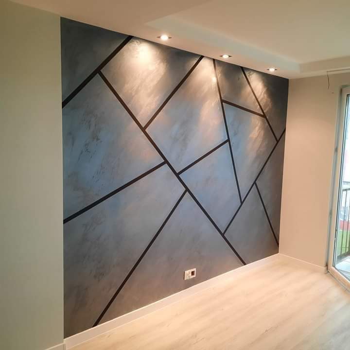 Ściana z dekoracyjną szarą strukturą i geometrycznym wzorem czarnych linii, oświetlona punktowymi lampami sufitowymi, widoczny fragment parkietu i okna balkonowego.