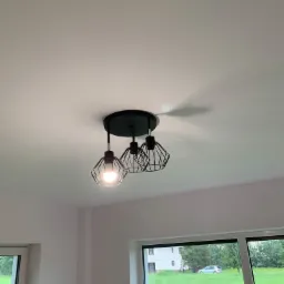 Czarny, nowoczesny plafon z trzema drucianymi kloszami, zamontowany na białym suficie w nowym wnętrzu z widokiem na zewnątrz przez okno.