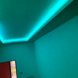 Pokój z podwieszonym sufitem i oświetleniem LED w kolorze turkusowym, widoczne rusztowanie i fragment okna z widokiem na dom.