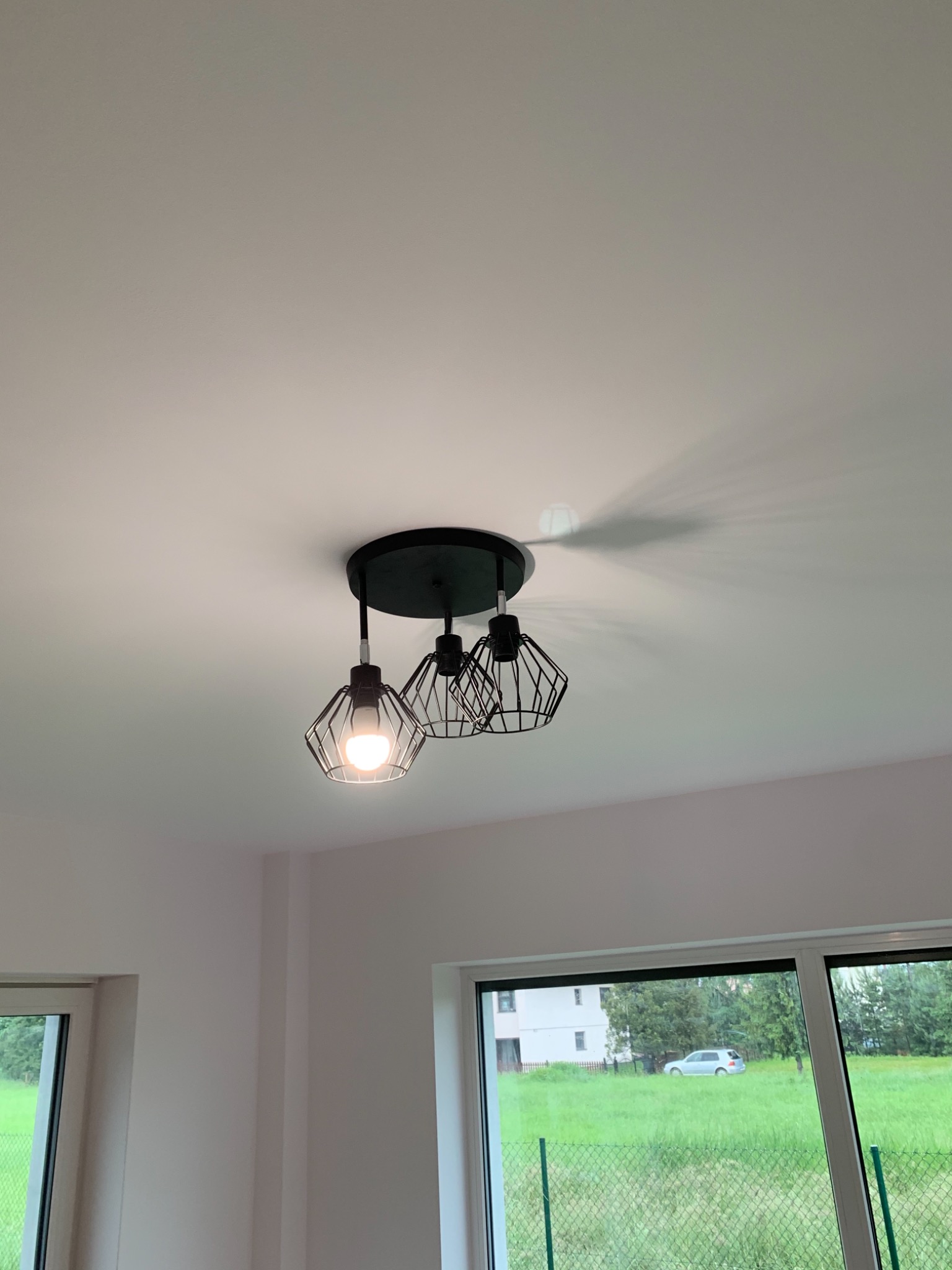 Czarny, nowoczesny plafon z trzema drucianymi kloszami, zamontowany na białym suficie w nowym wnętrzu z widokiem na zewnątrz przez okno.