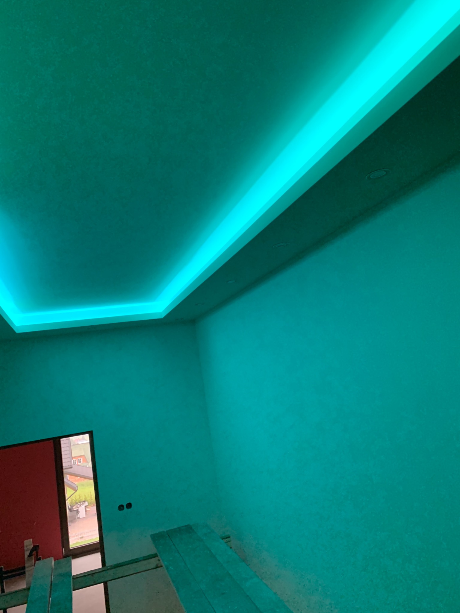 Pokój z podwieszonym sufitem i oświetleniem LED w kolorze turkusowym, widoczne rusztowanie i fragment okna z widokiem na dom.