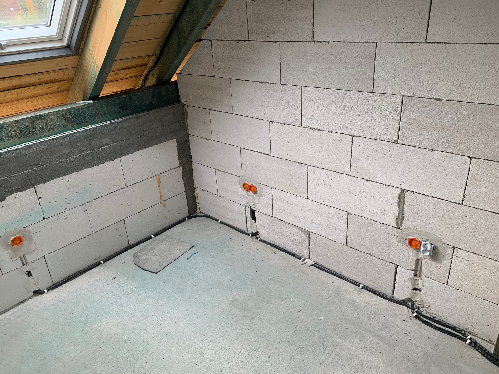 Surowy stan poddasza z widoczną instalacją elektryczną w peszlu, gniazdka w puszkach, ściany z betonu komórkowego, okno dachowe i drewniana konstrukcja dachu.