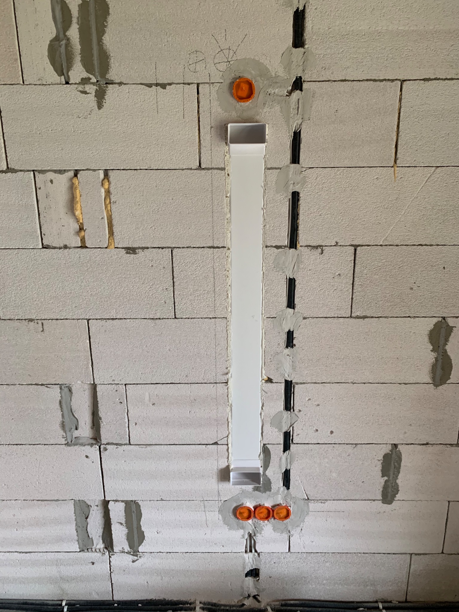 Ściana z betonu komórkowego z wyżłobionymi kanałami na przewody elektryczne, częściowo zakryte białą listwą maskującą i pomarańczowymi puszkami instalacyjnymi, z widocznymi przewodami w dolnej...