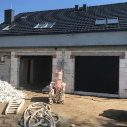 Budynek w trakcie budowy z białych bloczków, widoczna maszyna do tynkowania, sterta gruzu i zwinięte węże na placu budowy.