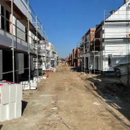 Budowa szeregu domów jednorodzinnych: widok wzdłuż ulicy z domami w trakcie ocieplania elewacji styropianem i murowania ścian z czerwonej cegły, z rusztowaniami i kontenerem na gruz w tle.