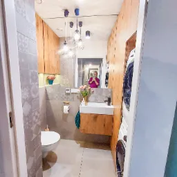 Wąska łazienka z toaletą, umywalką na drewnianej szafce, pralką w zabudowie i lustrem, w którym odbija się osoba robiąca zdjęcie.