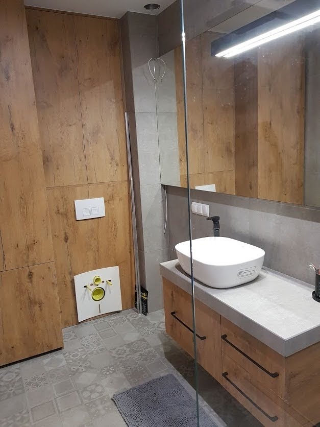 Nowoczesna łazienka z drewnopodobnymi panelami ściennymi, owalną umywalką nablatową i szarą mozaikową podłogą. Widoczny fragment kabiny prysznicowej i instalacja podtynkowa WC.