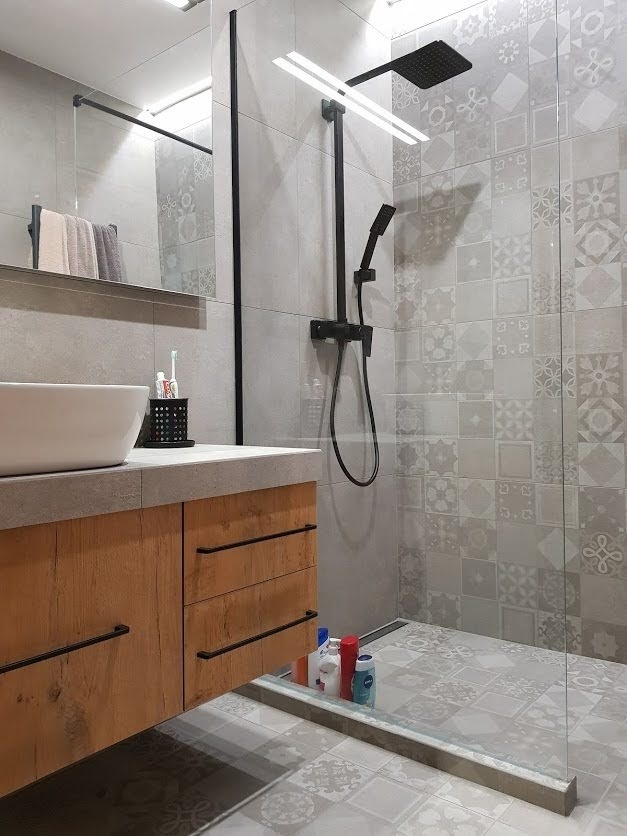 Nowoczesna łazienka z kabiną prysznicową typu walk-in, wykończona szarymi płytkami w patchworkowy wzór, drewniana szafka pod umywalką z czarnymi uchwytami, widoczne kosmetyki pod szafką.