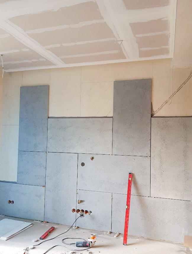 Montaż szarej okładziny ściennej z widocznymi otworami na gniazdka elektryczne i poziomnicą, sufit w trakcie wykończenia, widoczne narzędzia na podłodze.