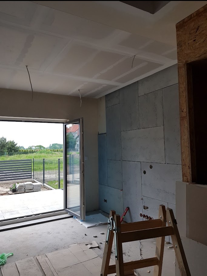 Wnętrze w trakcie prac wykończeniowych: ściana wyłożona płytami imitującymi beton, widoczne otwory na instalacje, sufit podwieszany z płyt gipsowo-kartonowych, rozsuwane drzwi balkonowe i drabina...