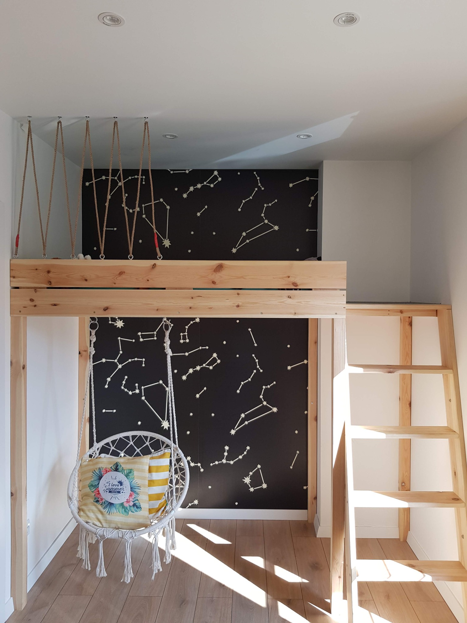 Pokój dziecięcy z drewnianym łóżkiem na antresoli, wiszącym krzesłem z poduszkami i ścianą w gwiazdy, oświetlony punktowymi lampami.