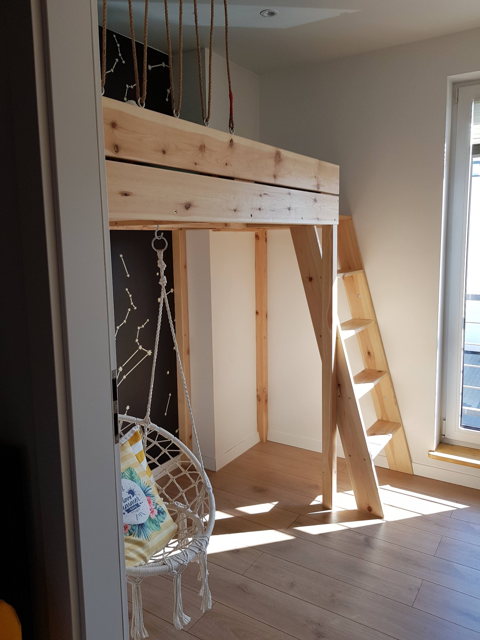 Pokój dziecięcy z drewnianym łóżkiem na antresoli, wiszącym fotelem i ścianą z motywem gwiazd, oświetlony naturalnym światłem.