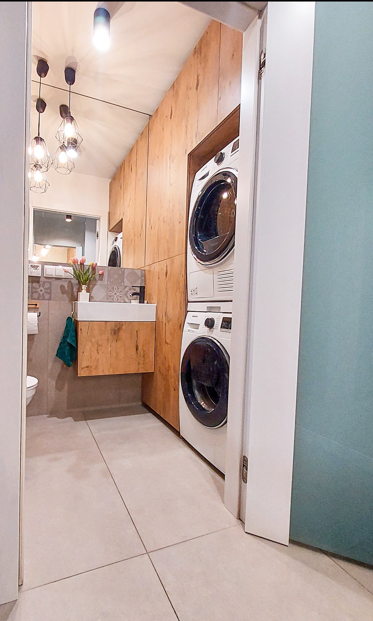 Nowoczesna łazienka z pralką i suszarką w zabudowie z drewnopodobnych paneli, wisząca szafka z umywalką, szara płytka na ścianie i podłodze, dekoracyjne lampy wiszące.