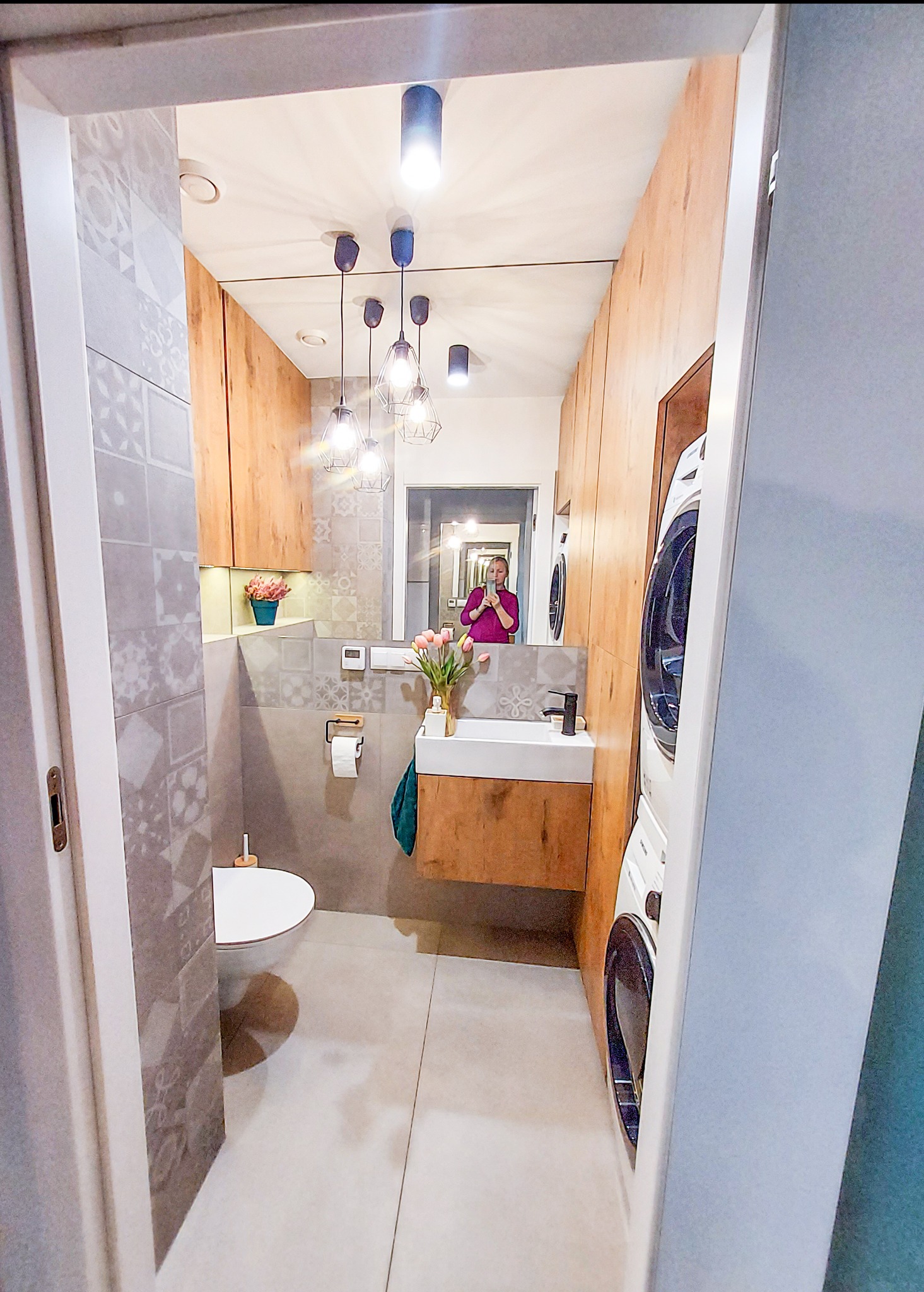 Wąska łazienka z toaletą, umywalką na drewnianej szafce, pralką w zabudowie i lustrem, w którym odbija się osoba robiąca zdjęcie.