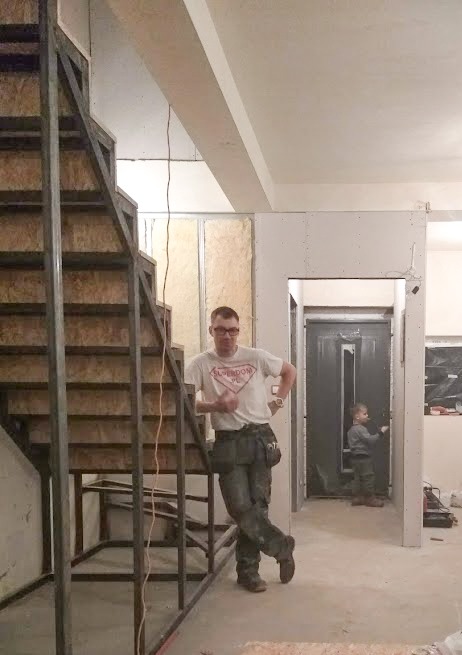 Pracownik w Żorach opiera się o metalową konstrukcję schodów w trakcie prac wykończeniowych wnętrza, w tle widoczna ściana z płyt kartonowo-gipsowych i małe dziecko.