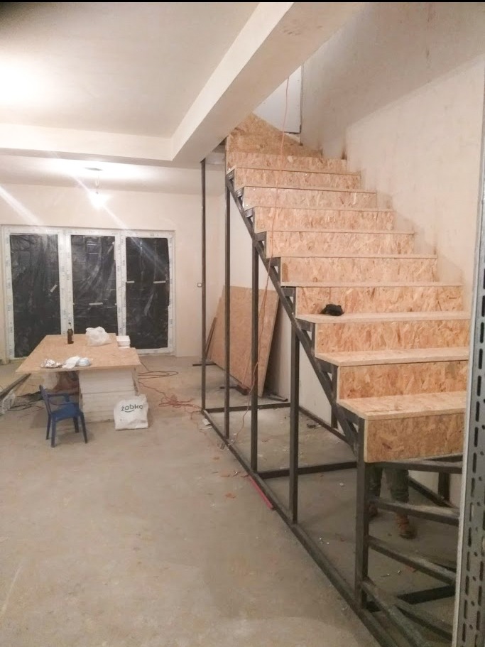 Wnętrze w trakcie remontu: surowe schody z metalową konstrukcją i płytami OSB, zabezpieczone okna folią, widoczny stół roboczy z materiałami budowlanymi i krzesłem, oraz worek z zaprawą.