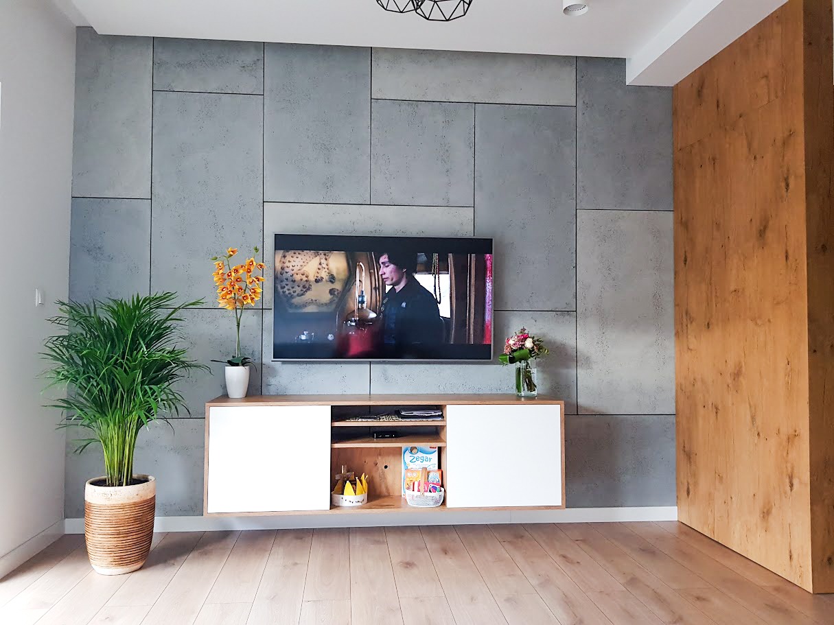 Nowoczesny salon z szarą ścianą imitującą betonowe płyty, wisząca szafka RTV z białymi frontami, drewniana okładzina ściany po prawej stronie i parkiet na podłodze.