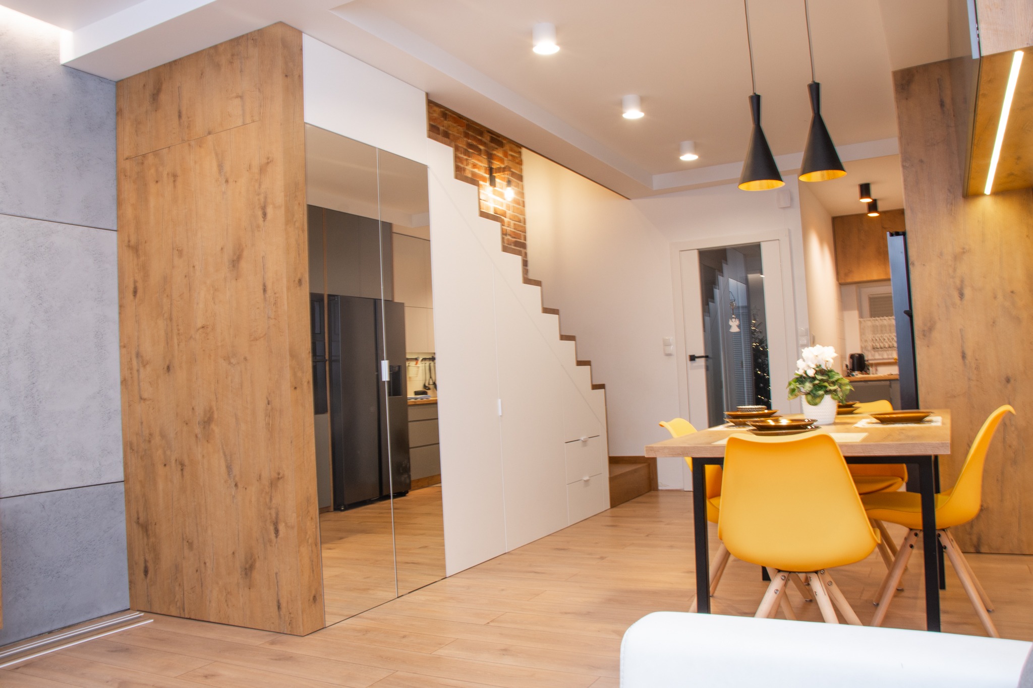 Jasne wnętrze mieszkania z drewnianą podłogą, żółtymi krzesłami przy stole, schodami z ceglanym akcentem i lustrzaną szafą w zabudowie, odbijającą kuchnię.
