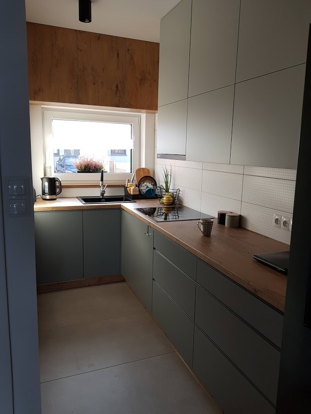 Nowoczesna kuchnia z szarymi szafkami, drewnianym blatem i panelami ściennymi imitującymi drewno, w tle widoczne okno z kwiatami.